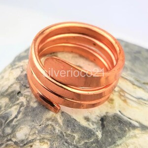 May include: A copper-colored spiral ring with a smooth, polished finish. The ring is coiled in a spiral shape, with multiple bands creating a layered effect. The ring is resting on a textured, gray and white stone surface.