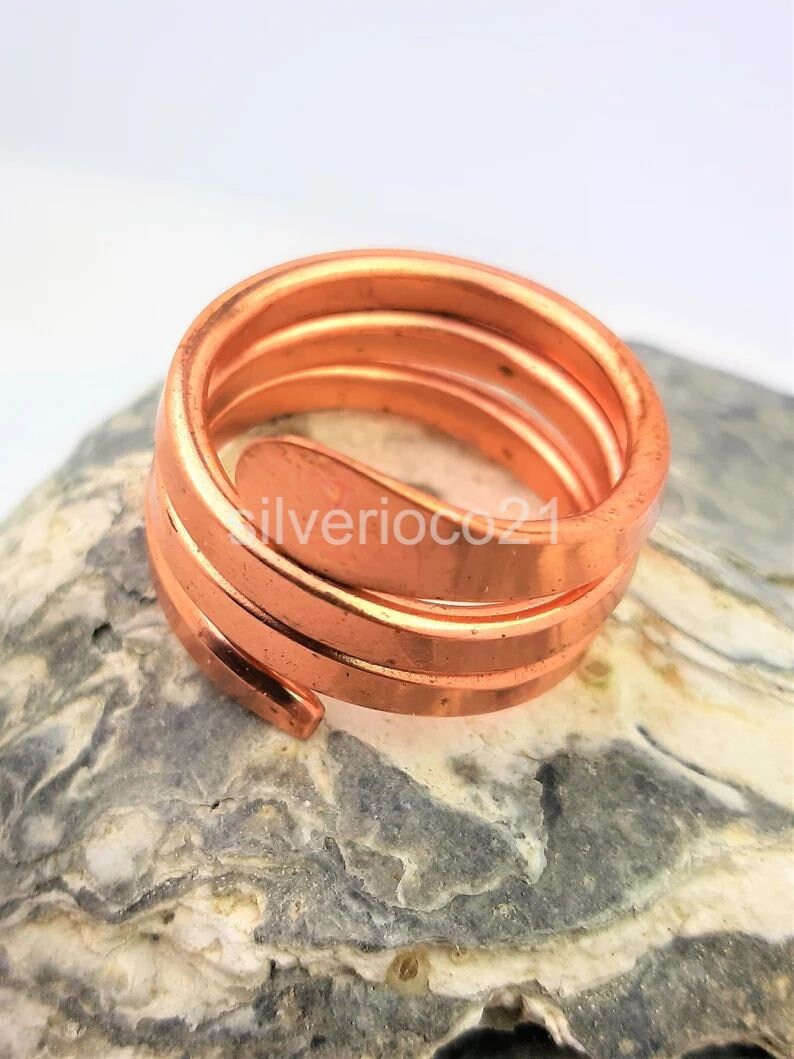 May include: A copper-colored spiral ring with a smooth, polished finish. The ring is coiled in a spiral shape, with multiple bands creating a layered effect. The ring is resting on a textured, gray and white stone surface.