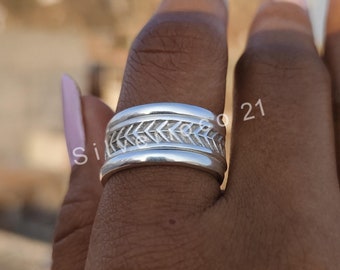 Könnte beinhalten: Ein silberner Ring mit einem detaillierten Fischgrätmuster. Der Ring ist breit und hat eine polierte Oberfläche. Das Design ist auf dem Ringband zentriert und schafft ein auffälliges Schmuckstück. Der Ring wird an einem Finger gezeigt.