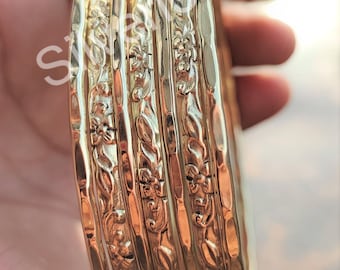 Puede incluir: Un conjunto de seis brazaletes de metal dorado con intrincados diseños florales. Los brazaletes están apilados y tienen una textura martillada.