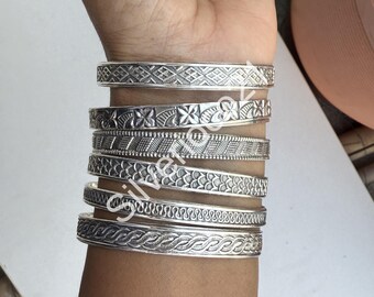 Pode incluir: Um conjunto de sete pulseiras de prata com designs intrincados. As pulseiras estão empilhadas no pulso de uma pessoa.