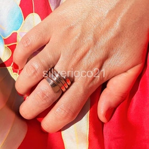 May include: A silver ring with a layered design is worn on a finger. The ring has a polished finish and a wide band. The background is a red and floral patterned fabric. The text "silverioco21" is visible.