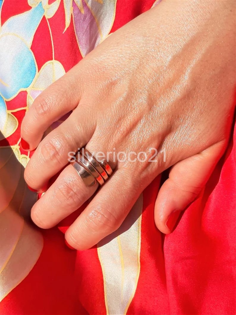 May include: A silver ring with a layered design is worn on a finger. The ring has a polished finish and a wide band. The background is a red and floral patterned fabric. The text "silverioco21" is visible.