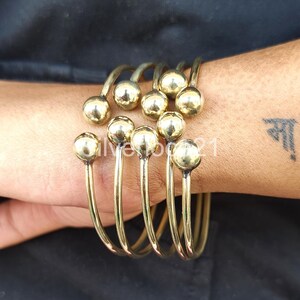 Pode incluir: Um conjunto de pulseiras de metal dourado. Cada pulseira apresenta um design em espiral com uma bola na extremidade de cada segmento curvo. As pulseiras são empilhadas em um pulso, mostrando seu design e textura. As pulseiras são de cor dourada quente.