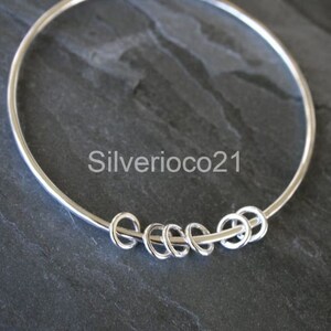 May include: A silver bangle bracelet with five small, interconnected circles. The bracelet is circular and has a smooth, polished finish. The text "Silverioco21" is visible on the bracelet.