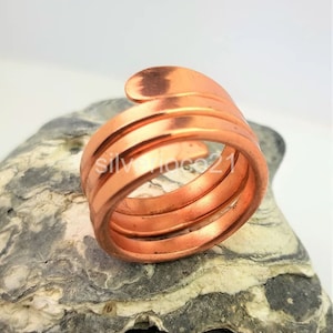 May include: A copper-colored ring with a spiral design. The ring wraps around the finger multiple times, creating a layered effect. The ring is made of a shiny metal and is displayed on a textured gray stone.