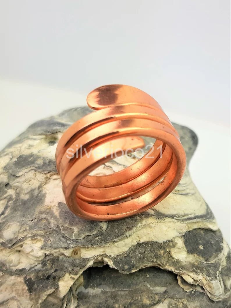 May include: A copper-colored ring with a spiral design. The ring wraps around the finger multiple times, creating a layered effect. The ring is made of a shiny metal and is displayed on a textured gray stone.