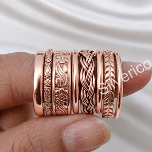 Könnte beinhalten: Ein Set aus acht roségoldfarbenen Ringen mit verschiedenen Designs, darunter ein glattes Band, ein geometrisches Muster, ein Blumenmotiv, eine geflochtene Textur und ein blattartiges Muster. Die Ringe sind nebeneinander an einem Finger angeordnet.