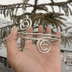 May include: A silver arm cuff with a spiral design at each end. The cuff is held in a hand, with the background showing greenery and a glimpse of a building. The word "Silvercoco" is visible on the cuff.