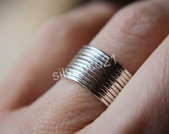 Könnte beinhalten: Nahaufnahme eines silbernen Rings mit einem strukturierten, geschichteten Design. Der Ring wird an einem Finger getragen und zeigt seine mehreren Bänder und seine reflektierende Oberfläche. Das Design des Rings ist detailliert und modern.