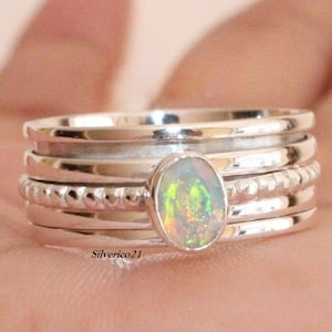 May include: A silver ring with a stack of five bands, featuring a green and white opal gemstone. The ring is on a person's finger.