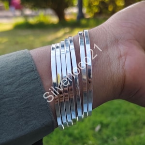 May include: A collection of shiny silver bangles stacked on a wrist. The bangles are flat and rectangular, reflecting light. The text "Silveroco21" is visible on the wrist. The person is wearing a green shirt.