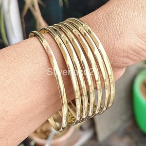 Op de afbeelding: Een set van zeven gouden metalen armbanden. De armbanden zijn op elkaar gestapeld en worden om de pols gedragen.