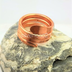 May include: A copper-colored spiral ring with a layered design. The ring is made of a continuous band of metal that wraps around the finger. The ring is displayed on a gray and white stone surface.