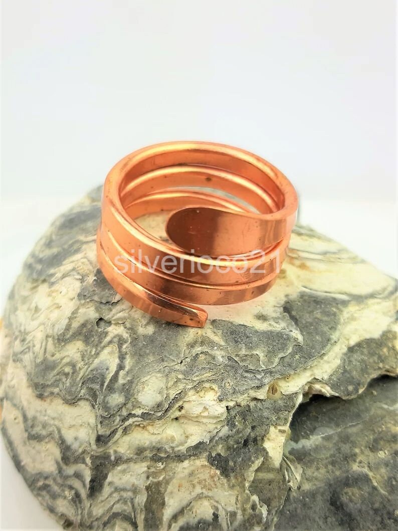 May include: A copper-colored spiral ring with a layered design. The ring is made of a continuous band of metal that wraps around the finger. The ring is displayed on a gray and white stone surface.