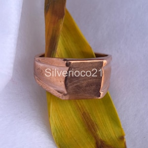 May include: A copper-coloured ring with a square top and a bevelled edge. The ring is engraved with the text "Silverico21".