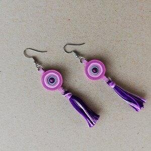 Puede incluir: Un par de pendientes de color rosa y morado con un borla. Los pendientes están hechos de papel y tienen un gancho de plata.