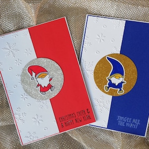 May include: Two Christmas cards with a white background and snowflake pattern. The card on the left has a red background with a silver glitter circle featuring a red gnome with a white beard. The text on the card reads "Christmas cheer & a happy new year". The card on the right has a blue background with a gold glitter circle featuring a blue gnome with a white beard. The text on the card reads "Jingle all the way!"