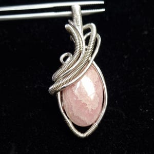 Könnte beinhalten: Handgefertigter Anhänger mit einem rosa Edelstein, der in eine Silberdrahtwicklung eingefasst ist. Der tropfenförmige Stein hat ein weiches, durchscheinendes Aussehen. Der Silberdraht ist kunstvoll um den Stein gewebt und schafft ein einzigartiges und elegantes Design.