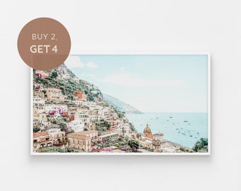 May include: A framed print of a coastal Italian town. The image features colorful buildings cascading down a hillside towards the sea. The sky is a soft blue, and the water is dotted with boats. A circular graphic in the upper left corner reads "BUY 2, GET 4".