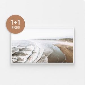 May include: An aerial photograph of a sandy beach with white waves crashing on the shore. The beach is bordered by a line of trees and a hazy sky.