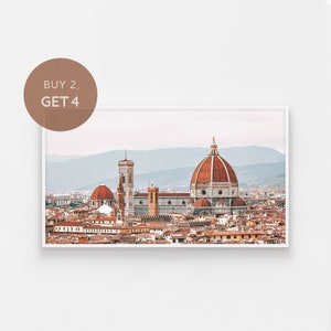 May include: A panoramic view of the city of Florence, Italy, featuring the iconic Duomo cathedral in the centre. The cathedral has a large, red-brown dome and is surrounded by other buildings, including a tall bell tower. The city is bathed in a soft, warm light.