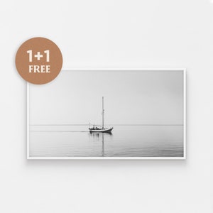 May include: A black and white photograph of a sailboat on a calm body of water. The sailboat is in the centre of the image and is facing the right side of the frame. The water is smooth and there is a faint reflection of the sailboat in the water.