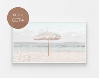May include: A white beach umbrella with a fringe border stands on a sandy beach with a blue ocean in the background.
