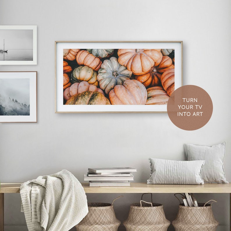 May include: A framed print of a close-up of a pile of colorful pumpkins. The print is hanging on a white wall above a wooden shelf with books, a blanket, and baskets.