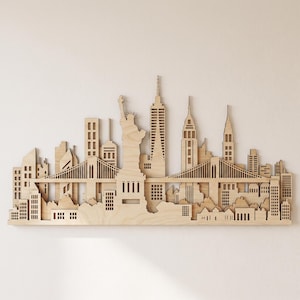 May include: A laser-cut wooden wall art piece depicting the New York City skyline. The design features the Statue of Liberty, skyscrapers, and suspension bridges layered to create depth. The natural light wood grain is visible throughout the structure.