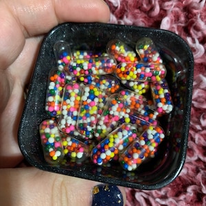 Puede incluir: Un bol negro lleno de c&aacute;psulas transparentes que contienen coloridos sprinkles. El bol est&aacute; sostenido en una mano con esmalte de u&ntilde;as azul oscuro con brillo dorado.