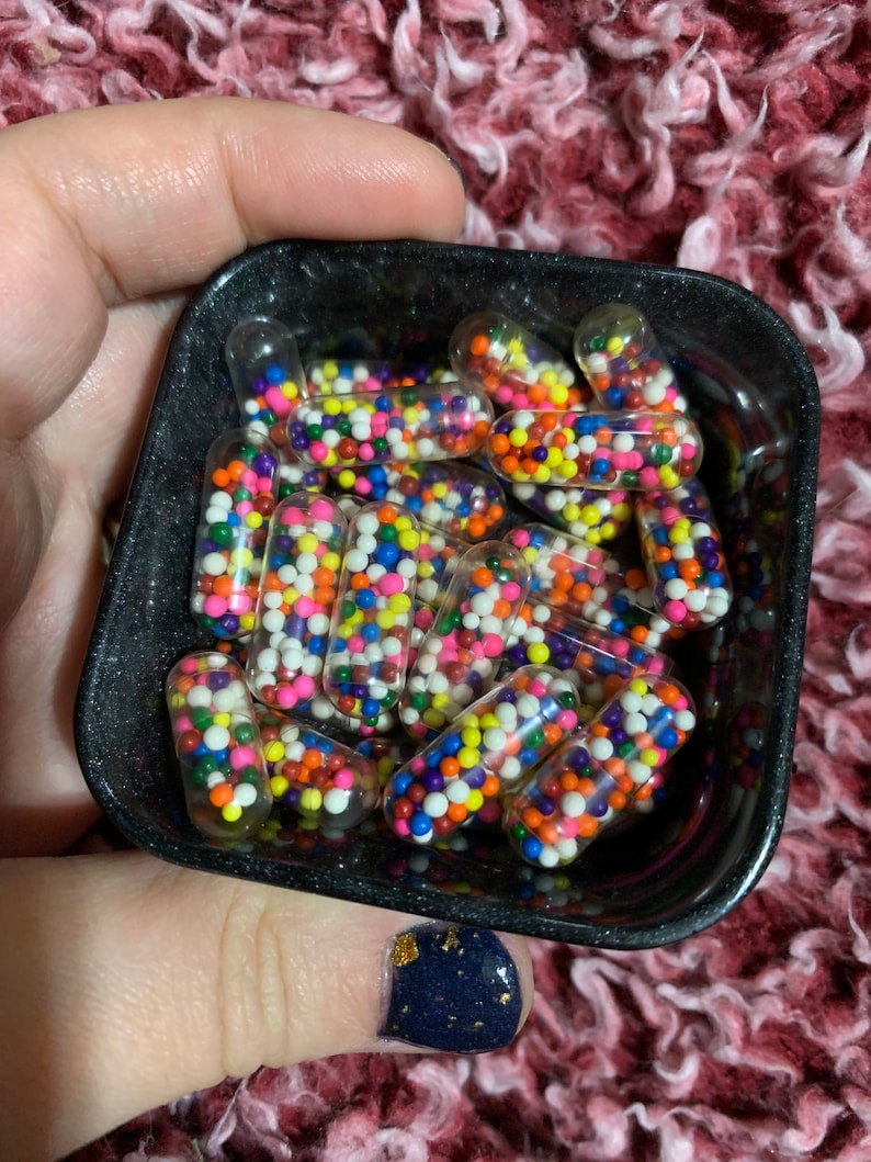 Puede incluir: Un bol negro lleno de c&aacute;psulas transparentes que contienen coloridos sprinkles. El bol est&aacute; sostenido en una mano con esmalte de u&ntilde;as azul oscuro con brillo dorado.