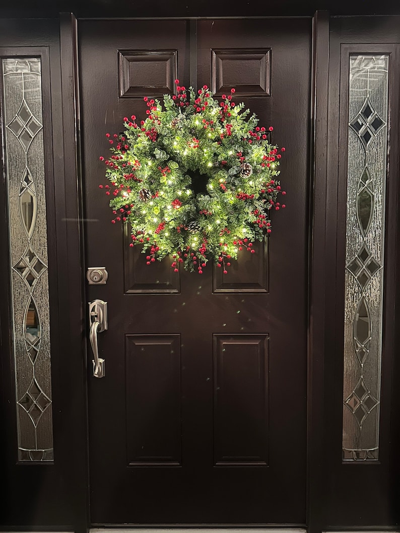 May include: A dark brown wooden door with a wreath hanging on it. The wreath is made of green leaves and red berries. There are small white lights on the wreath. The door has a silver door handle and a silver deadbolt lock.