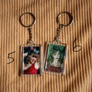 May include: Two keychains with rectangular photo inserts. Each keychain has a silver ring and chain. The photos feature a woman in a red dress and a woman with green hair. The background is a tan corduroy fabric.