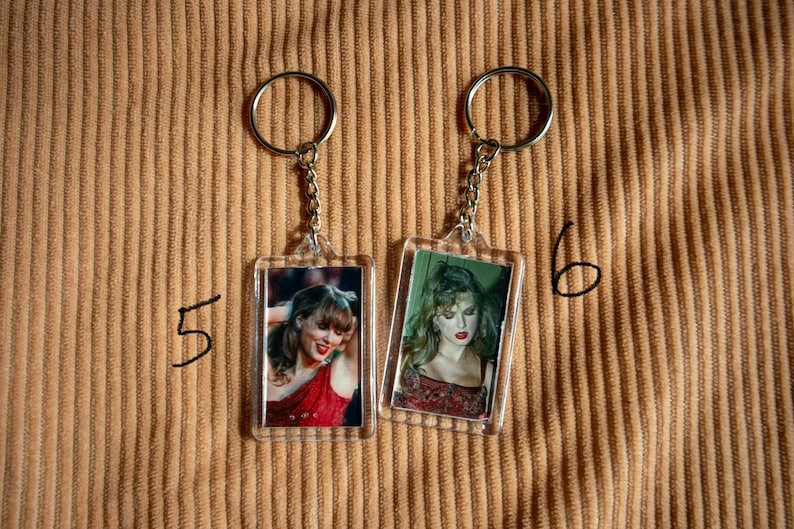 May include: Two keychains with rectangular photo inserts. Each keychain has a silver ring and chain. The photos feature a woman in a red dress and a woman with green hair. The background is a tan corduroy fabric.