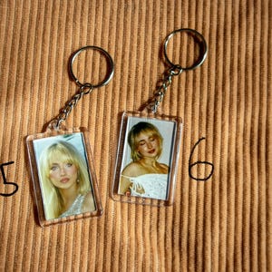 May include: Two clear acrylic keychains with silver chains and rings. Each keychain features a rectangular photo of a woman with blonde hair. The keychains are on a textured, tan surface. Numbers 5 and 6 are written in black marker.