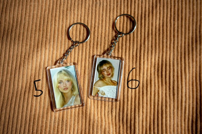 May include: Two clear acrylic keychains with silver chains and rings. Each keychain features a rectangular photo of a woman with blonde hair. The keychains are on a textured, tan surface. Numbers 5 and 6 are written in black marker.