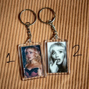 May include: Two keychains with rectangular photo inserts. One keychain features a color photo of a woman in a black and pink top. The other keychain has a black and white photo of a woman with her finger to her lips. Both have silver key rings.