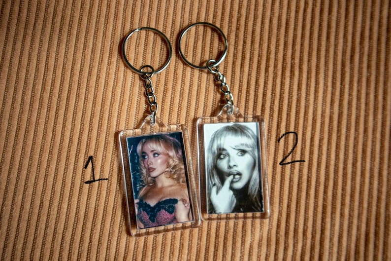 May include: Two keychains with rectangular photo inserts. One keychain features a color photo of a woman in a black and pink top. The other keychain has a black and white photo of a woman with her finger to her lips. Both have silver key rings.