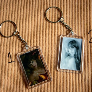 May include: Two rectangular keychains with metal rings and chains. One keychain features a color image of a woman in a red dress, the other a black and white photo of a woman. The background is a brown, corduroy-like fabric.