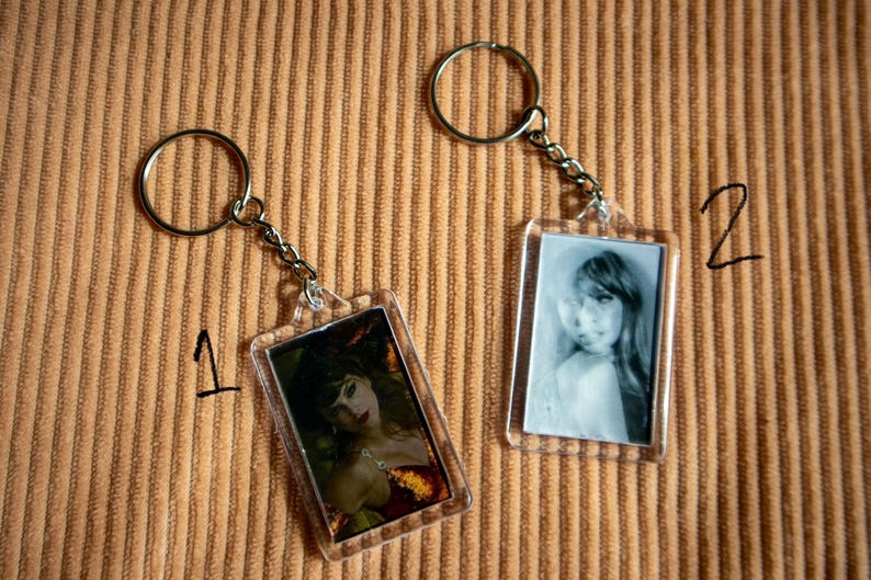 May include: Two rectangular keychains with metal rings and chains. One keychain features a color image of a woman in a red dress, the other a black and white photo of a woman. The background is a brown, corduroy-like fabric.