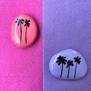 May include: Two painted rocks with palm tree silhouettes. One rock is pink with two palm trees and the other is purple with three palm trees.