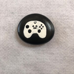 May include: A black painted rock with a white painted video game controller design. The controller has a white X button and a white circle button.
