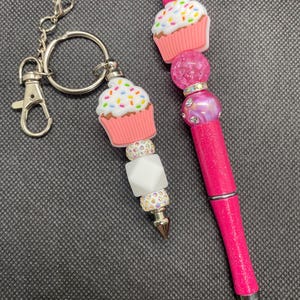 May include: A pink cupcake-themed keychain and pen set. The keychain features a cupcake charm with colorful sprinkles, white frosting, and a pink base. The pen has a matching cupcake topper and pink barrel with silver accents.