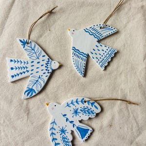 May include: Three white ceramic bird decorations with blue floral patterns and brown twine hangers. Each bird has a yellow beak and is decorated with different blue designs. The decorations are on a beige fabric surface.