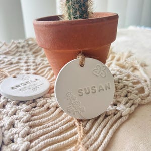 May include: A small cactus in a terracotta pot sits on a macrame placemat. Two white, round ceramic tags with the names "Angela" and "Susan" are visible. The "Susan" tag has floral and butterfly designs.