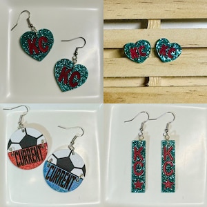 May include: A collection of four pairs of earrings. Two pairs are heart-shaped with teal glitter and the letters "KC" in red. One pair is round with a soccer ball design and the word "CURRENT". The last pair is rectangular with teal glitter, the letters "KC", and a red star.