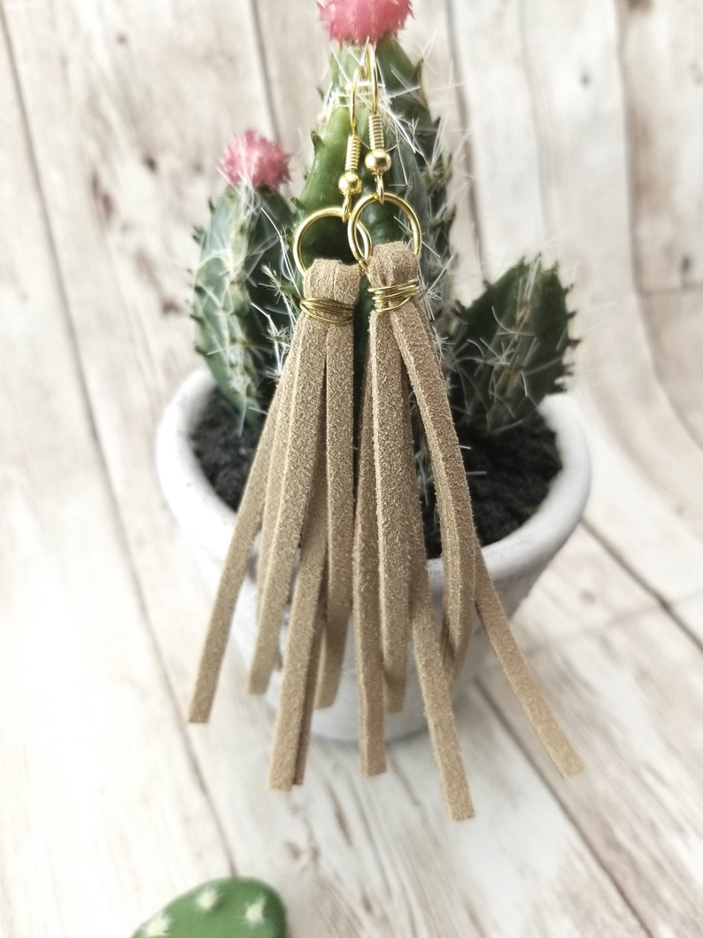 May include: A pair of brown suede tassel earrings with gold-toned hoops and wire wraps. The earrings are hanging from a potted cactus.