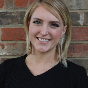 May include: A woman with blonde hair is smiling and wearing a black and brown sweater. She is standing in front of a brick wall.