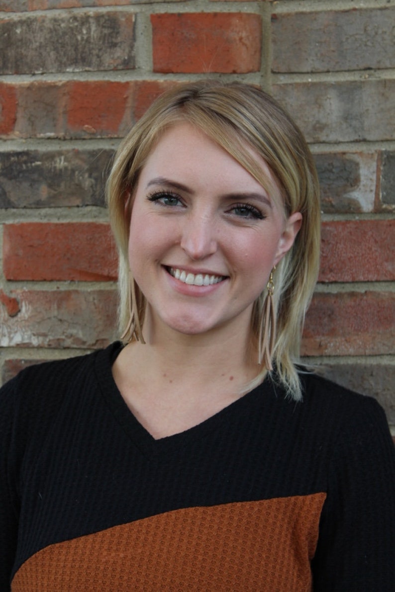 May include: A woman with blonde hair is smiling and wearing a black and brown sweater. She is standing in front of a brick wall.